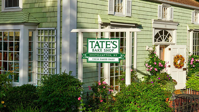 Front view of Tate's Bake Shop in Southampton, NY, with a green exterior, surrounded by flowers and a sign that reads "Tate's Bake Shop - Fresh Baked Daily.