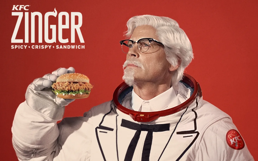 Colonel Sanders dressed as an astronaut, holding a KFC Zinger sandwich in a KFC commercial showcasing the iconic fast-food character.