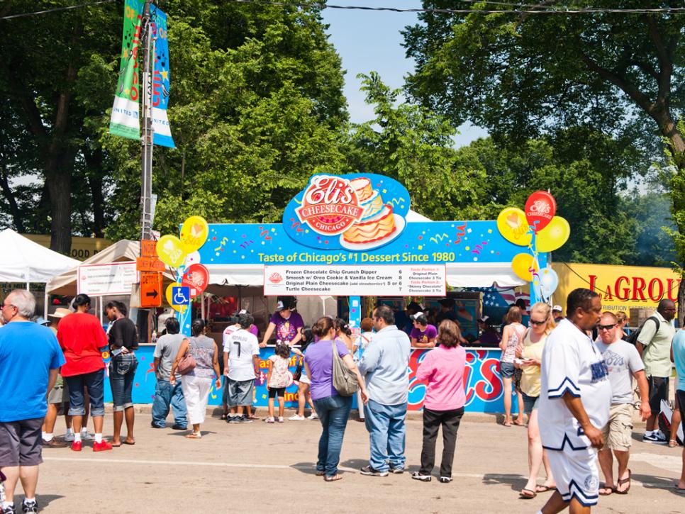 A crowded booth at a food festival promoting Eli's Cheesecake, labeled as "Chicago's #1 Dessert," with people gathering to purchase treats.